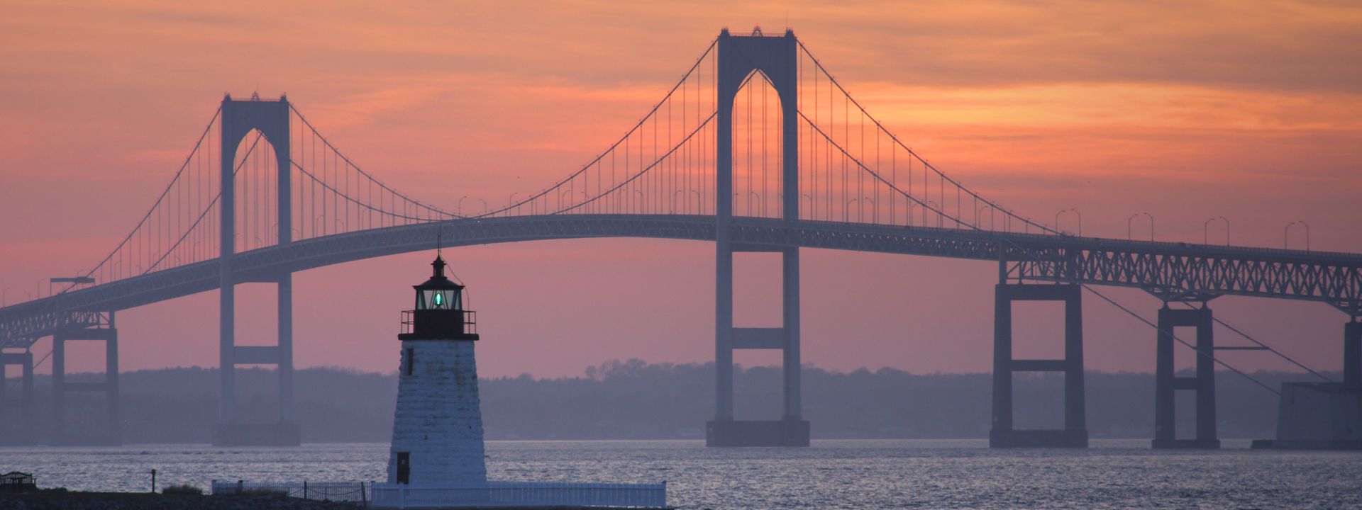 A lighthouse in front of the Claiborne Pell Bridge at sunset.