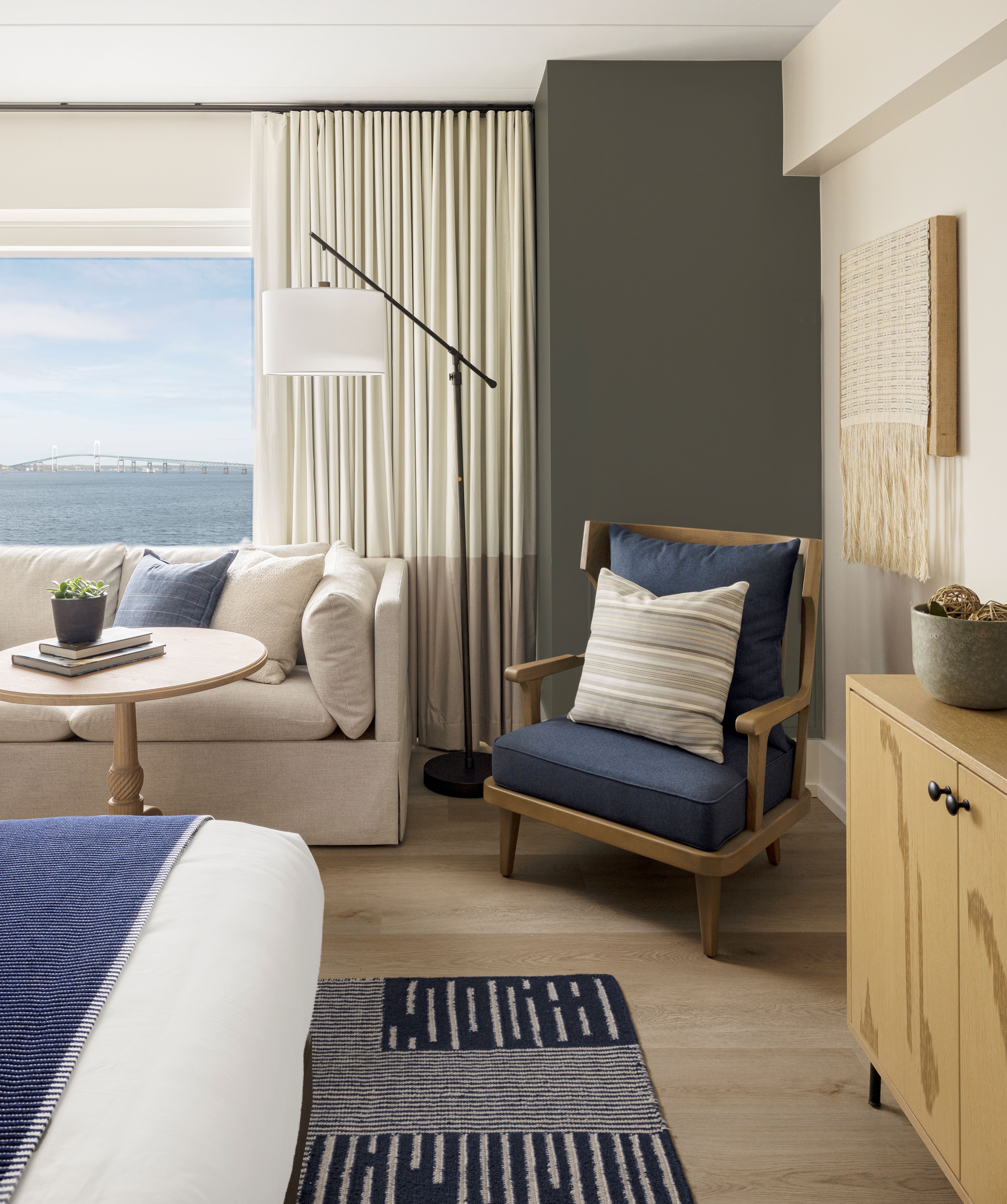 A furnished living room with a sofa, chair, coffee table, and lamp, with a large window showing an ocean view.