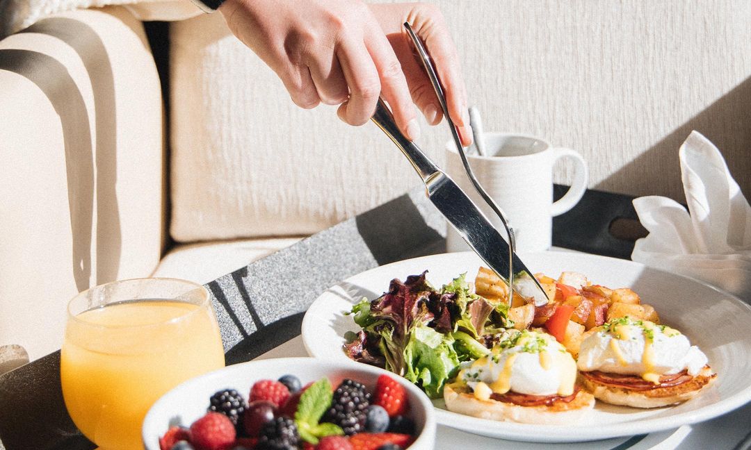 A person cutting into a plate of eggs benedict on a breakfast tray