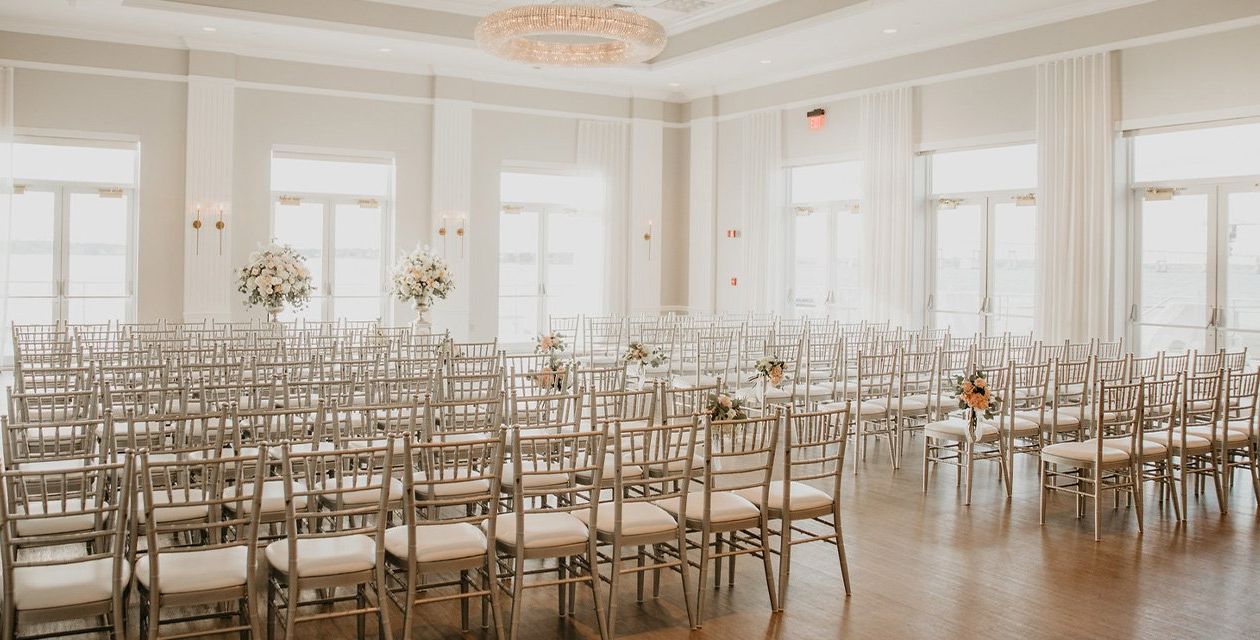 A white wedding venue with several windows set with rows of chairs.