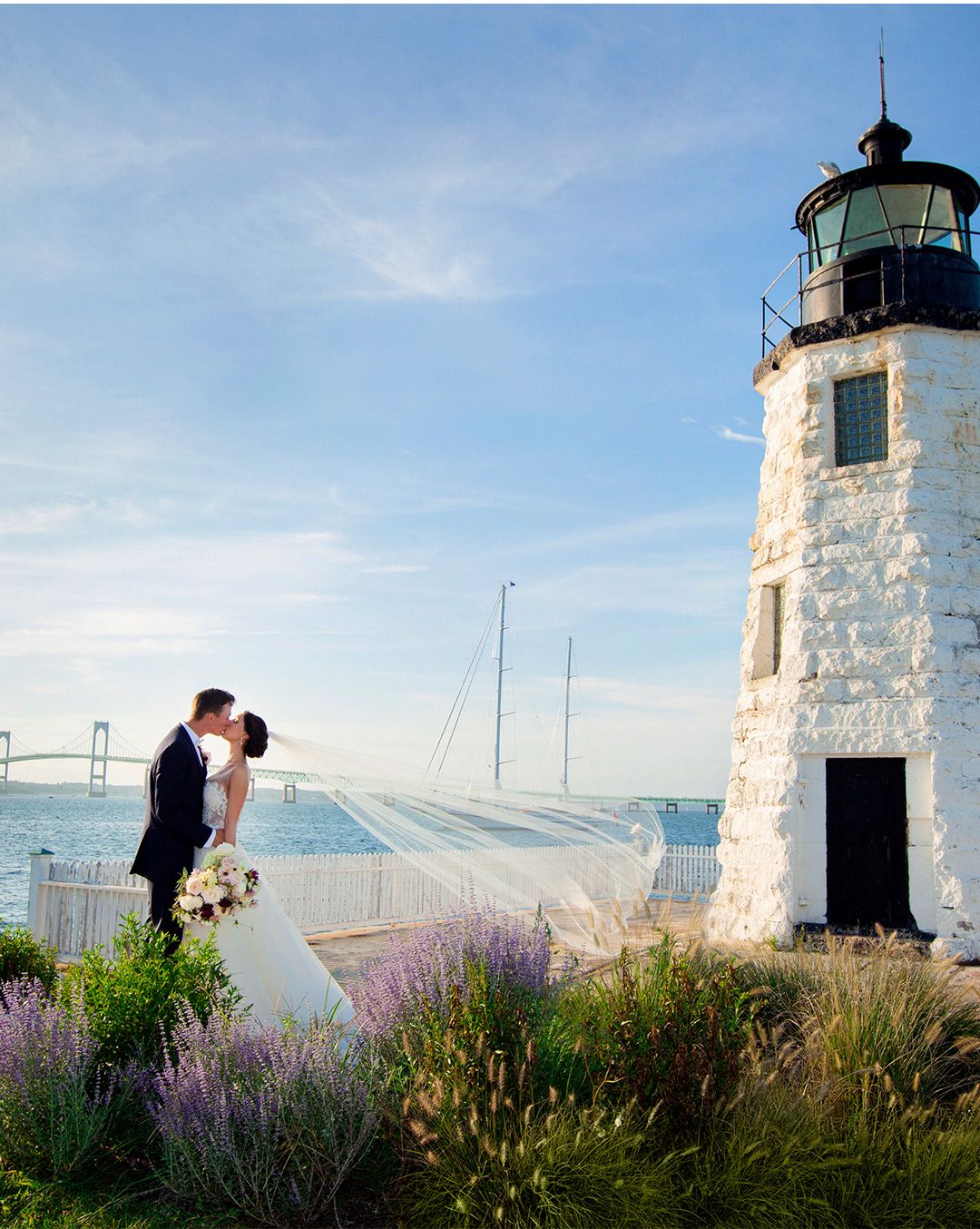 a white lighthouse next to a body of water with a bridge in the background and a couple in wedding attire