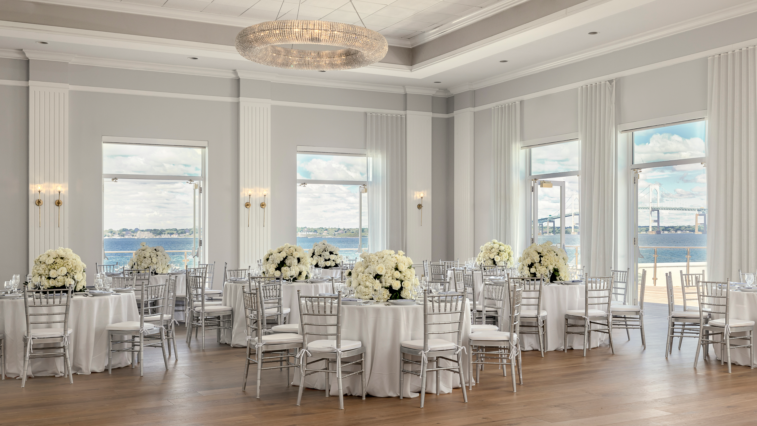 a brightly lit banquet hall with white tables and chairs, white flowers, and a large chandelier