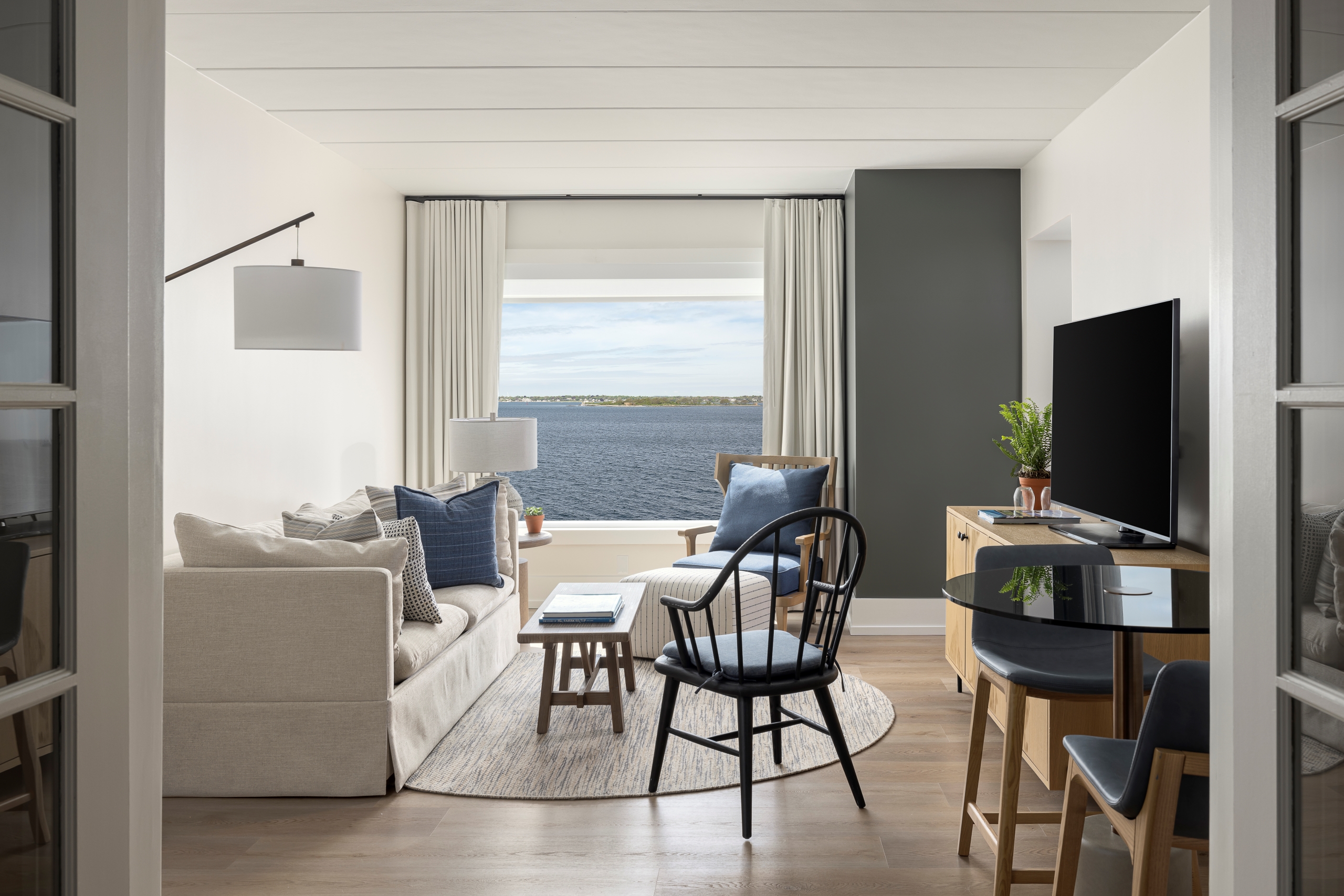 A bright living room with a sofa, chair, coffee table, and TV, with a sea view through the window.