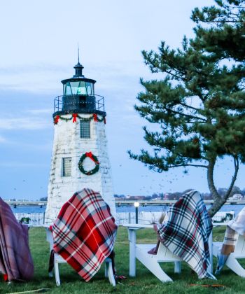 A white lighthouse with a black roof and a wreath on the front is shown with several plaid blankets on outdoor chairs in the foreground.