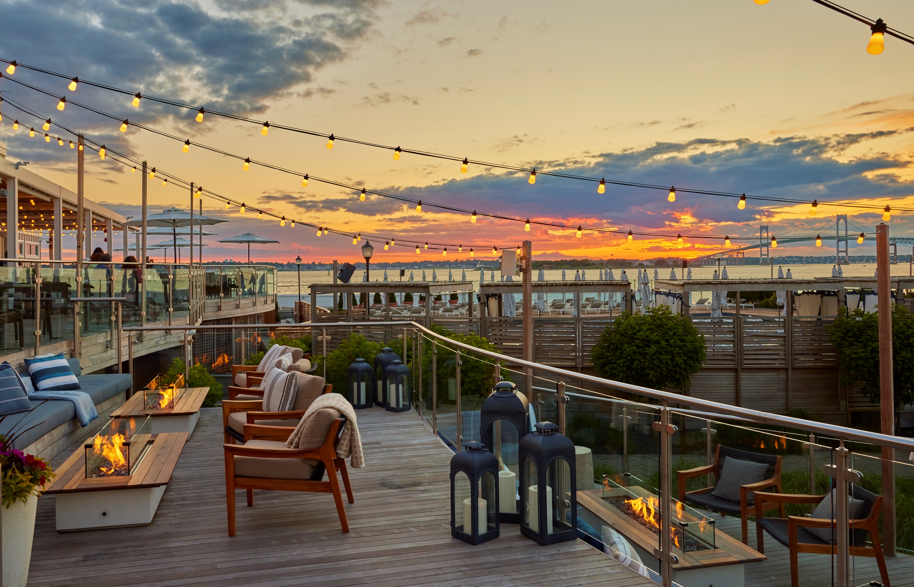 A rooftop patio with a sunset view of the water, outdoor seating, fire pits, and string lights.