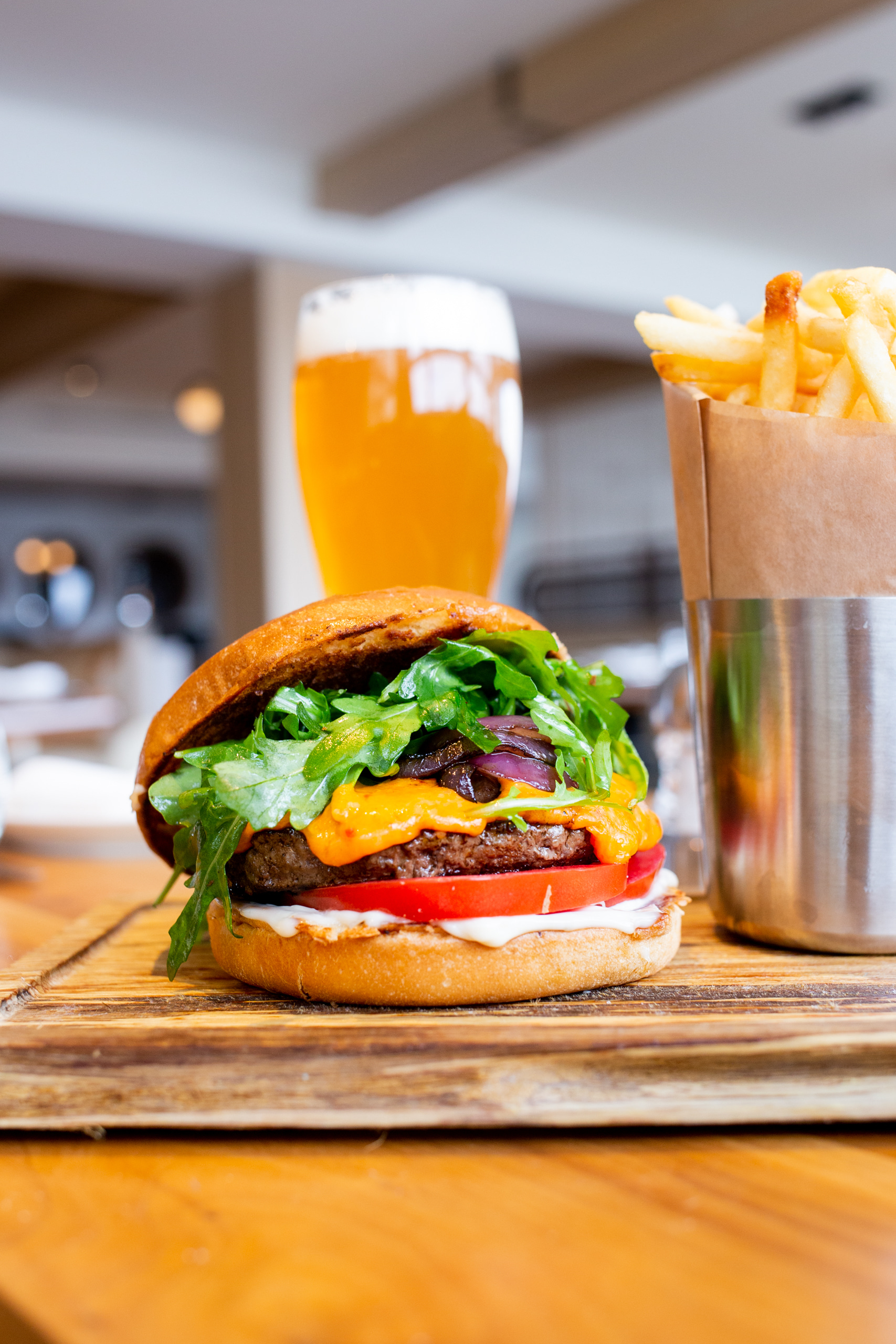 a burger with lettuce, tomato, and cheese on a wooden table with a metal cup of fries and a glass of beer in the background