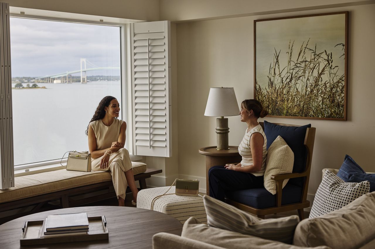 Two women sitting in a furnished room with a large window overlooking the water.