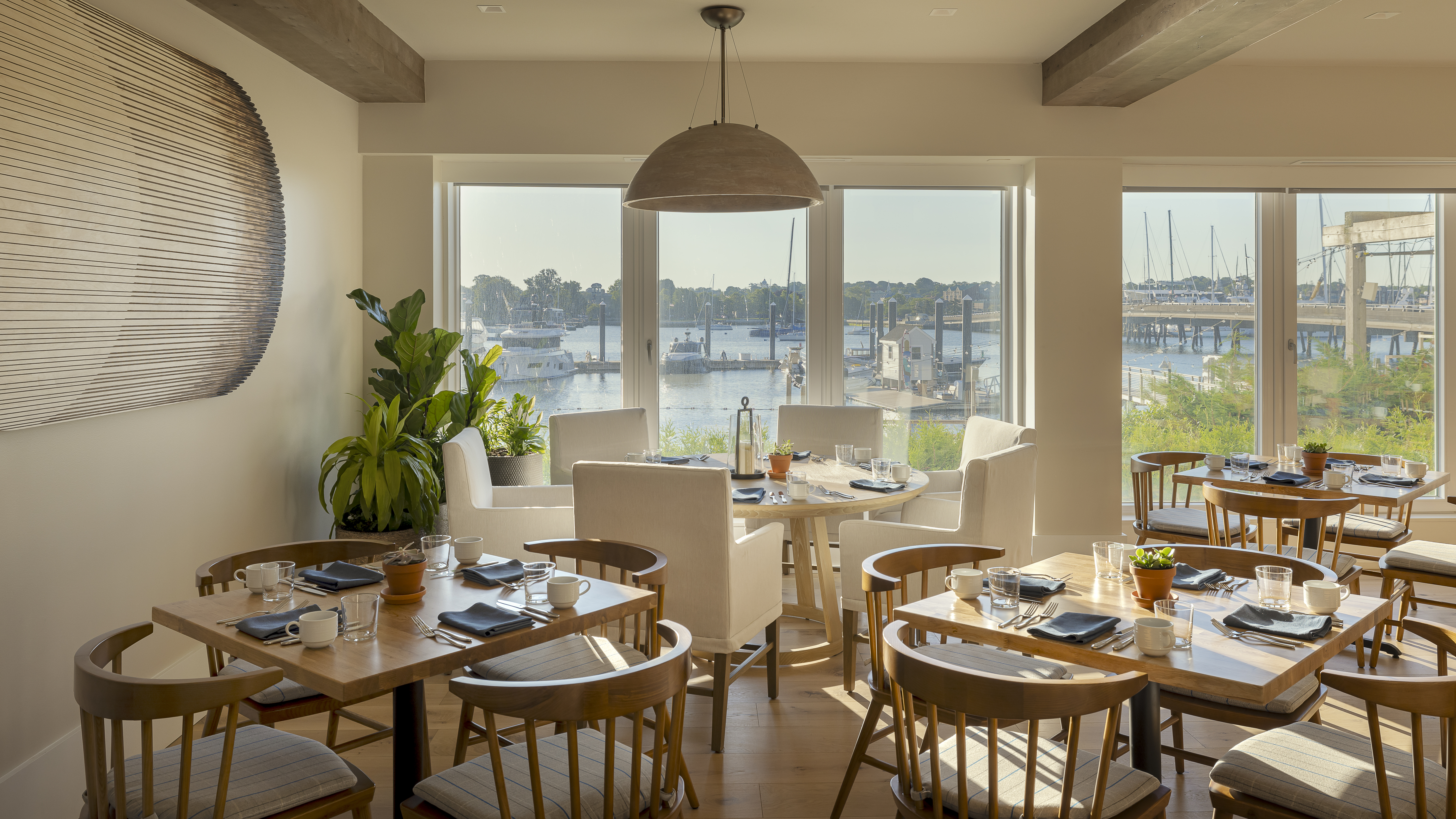 A dining room with several tables set for meals, large windows offering a view of the water and boats docked outside.