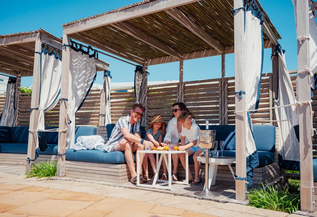 A family of four enjoys a meal in a cabana with blue couches and white curtains.
