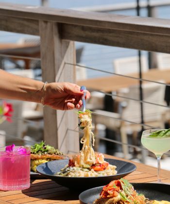 A hand is shown placing pasta on a plate with other dishes and drinks on a table with an outdoor setting.
