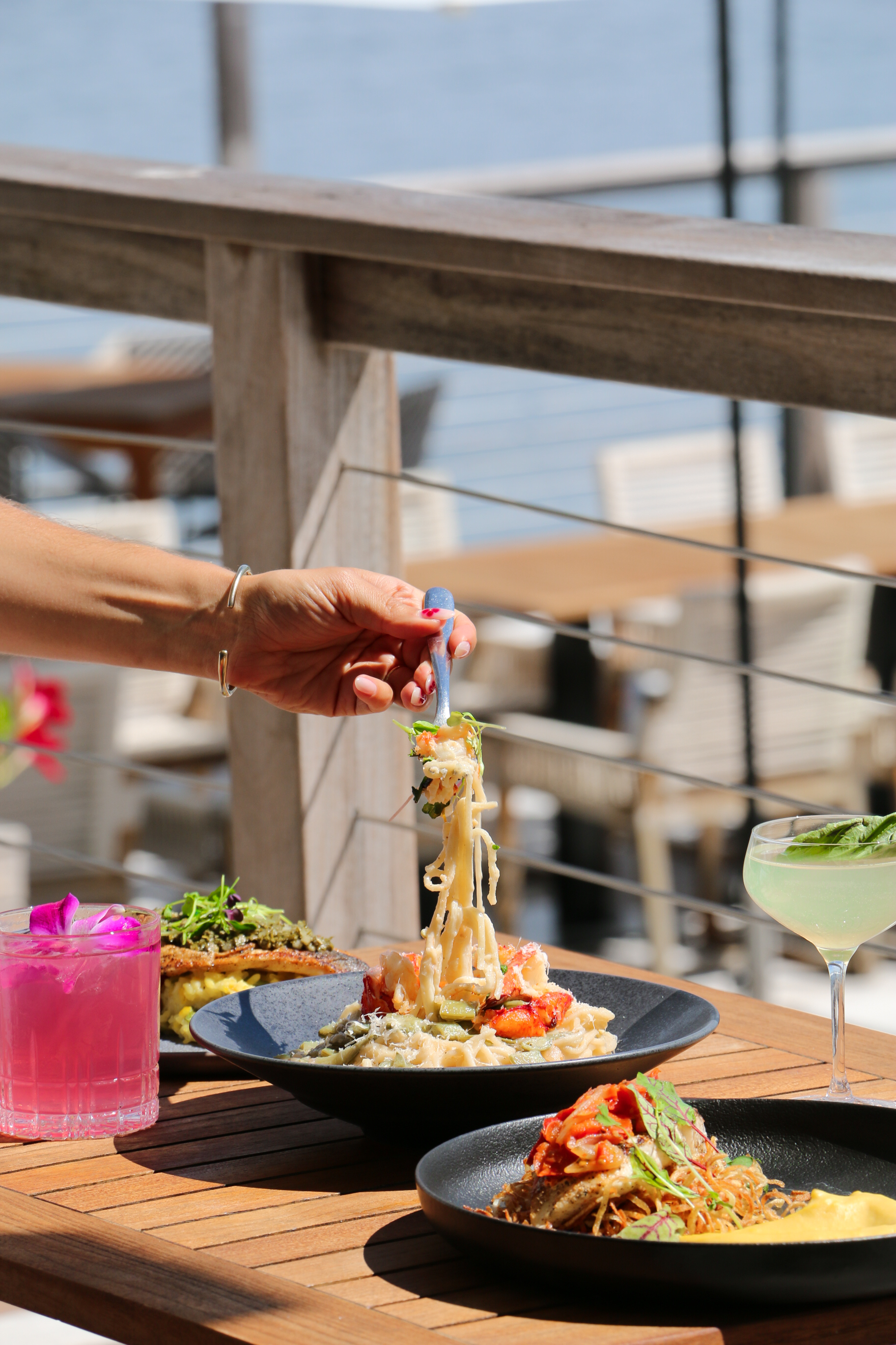 A hand is shown placing pasta on a plate with other dishes and drinks on a table with an outdoor setting.